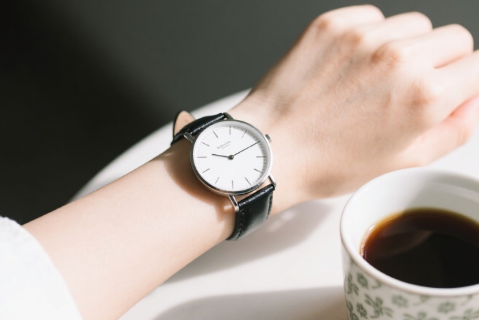 Thin minimalist analog watch with leather band on wrist near a white coffee mug